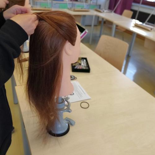 Eine Schülerin übt eine Flechtfrisur an einem Friseurpuppenkopf mit langen rotenbraunen Haaren