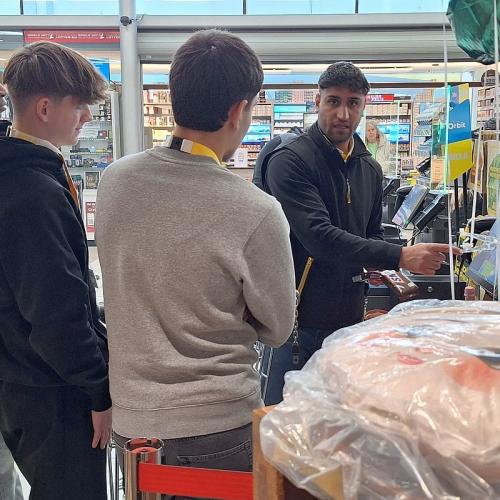 Bild zeigt denselben Kassenmitarbeiter wie 3 Schülern den Arbeitsablauf an einer Kassa zeigt