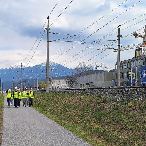 Bild zeigt eine Schülergruppe in gelben Warnwesten und weißen Bauhelmen auf einer Straße im Hintergrund die Zentrale der Firma Porr