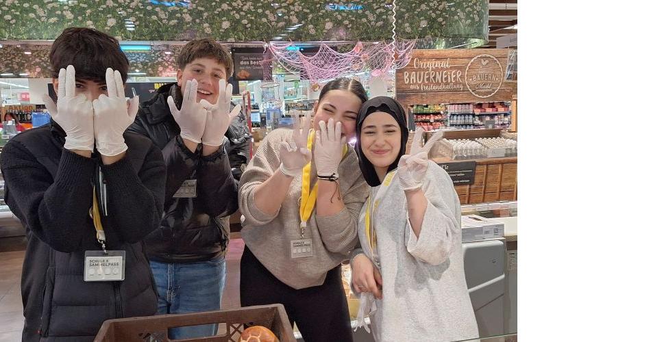Bild zeigt vier Schüler*innen in einer Billa-Filiale. Sie halten ihre Hände in die Kammera und tragen Einweghandschuhe.